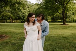 Bride smiling as groom leans in close outdoors, both in wedding attire on a lush green lawn.