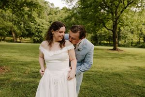 Groom in light blue suit whispers to laughing bride in off-shoulder wedding dress in a sunny green park.