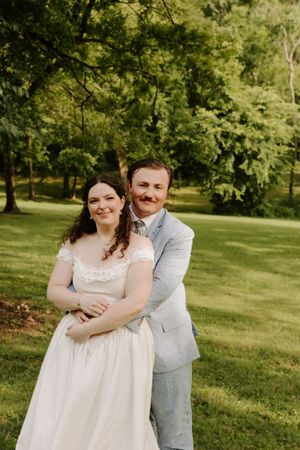 Groom embracing bride from behind on green lawn, both smiling at camera, bride in white gown and groom in blue suit.
