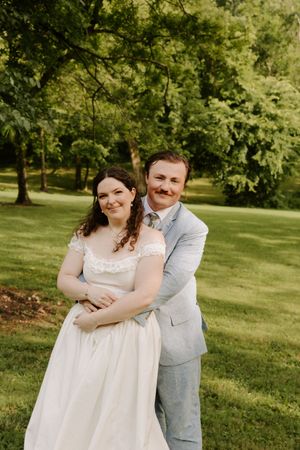 Wedding couple posing together outdoors under trees, groom in light blue suit embracing bride in white lace gown.