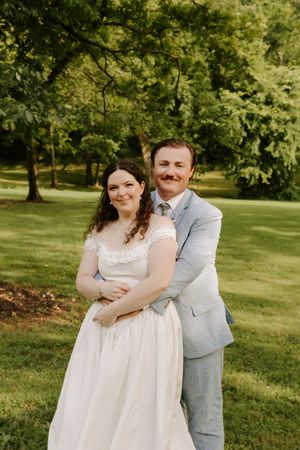 Smiling bride and groom pose together outdoors, groom embracing bride from behind in a sunny green park setting.