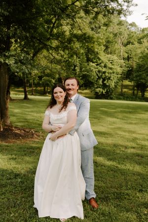 Wedding couple poses in sunlit garden, groom in light blue suit embracing bride in elegant white off-shoulder wedding dress.