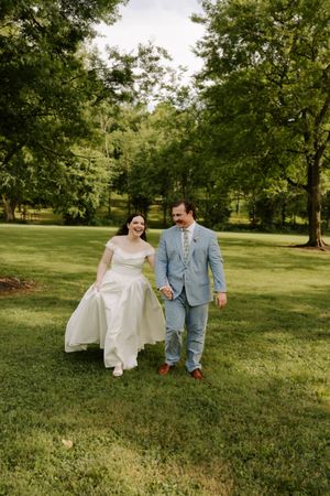 Bride and groom walk hand-in-hand through lush green park, bride lifting white gown, groom in light blue suit.