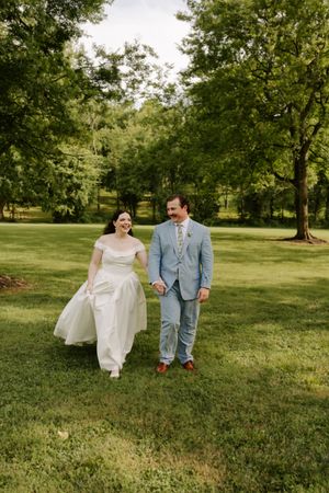 Happy couple strolls across green lawn on wedding day, bride in white off-shoulder gown, groom in blue suit.