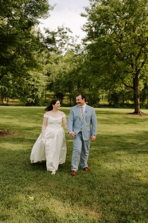 Bride in white off-shoulder gown and groom in blue suit walk hand-in-hand through a green park.