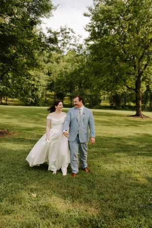 A joyful bride and groom walk hand in hand across a lush green lawn surrounded by tall trees.