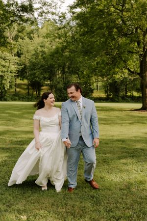 Smiling bride and groom walk together holding hands on a green lawn with tall trees in the background.