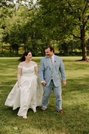 Joyful bride and groom walking hand-in-hand through green park, laughing together on their wedding day.