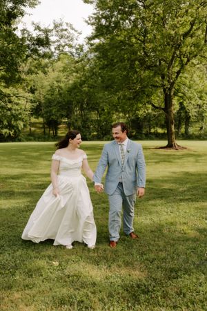 Bride in off-shoulder gown and groom in light blue suit walk hand-in-hand across green lawn at wedding.