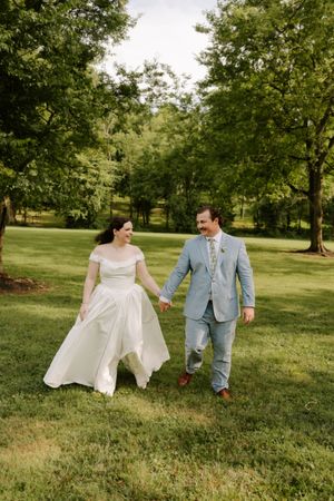 Joyful bride and groom walk hand-in-hand through a scenic green park, bride lifting her white gown while smiling at groom.