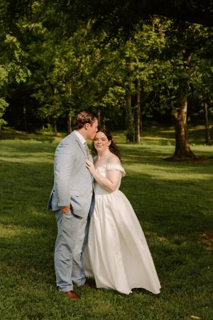 Groom tenderly kisses bride on cheek in golden sunlight among tall green trees on wedding day.