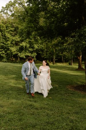 Bride and groom holding hands and walking joyfully across a green lawn surrounded by trees.