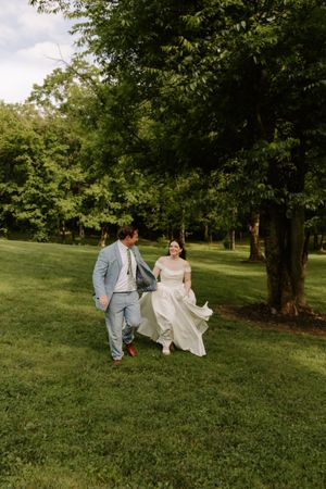 Bride and groom run hand in hand across green grass beneath tall trees.