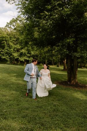 Wedding couple walks playfully through scenic green park, bride in flowing white dress, groom in light blue suit.