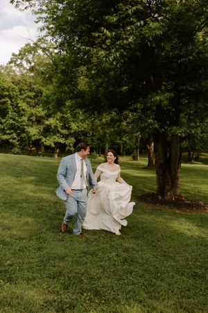 Joyful bride and groom run hand-in-hand across lush green lawn with tall trees in background.