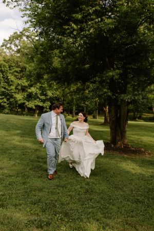 Joyful bride lifts white wedding gown while running with groom through a lush green outdoor venue.