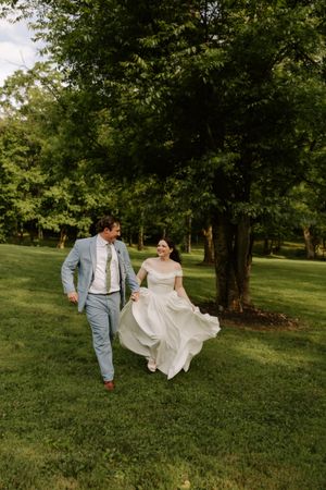 Smiling bride lifts white gown while walking with groom in light blue suit through green park.