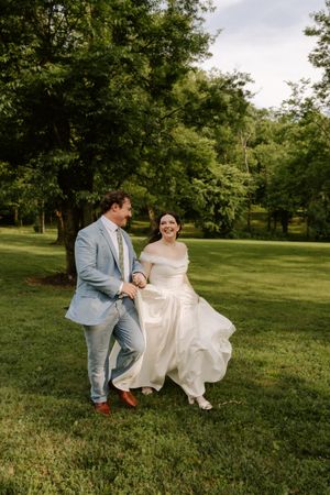 Joyful bride and groom walk hand-in-hand across green lawn, bride laughing in flowing white off-shoulder wedding gown.