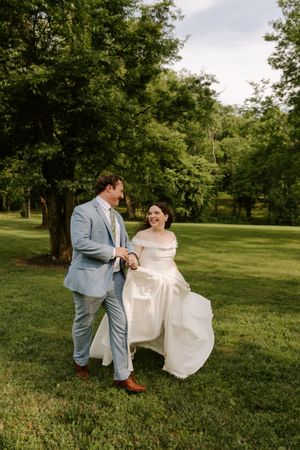 A happy bride lifts her white gown while walking hand-in-hand with her groom across a green lawn.