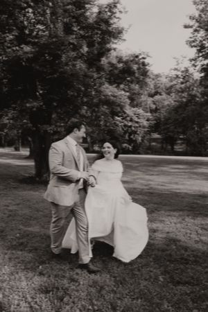Black and white photo of a bride and groom holding hands and laughing while walking through a lush green park.