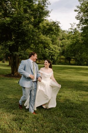 Happy bride and groom laugh together while strolling across green grass surrounded by lush trees.