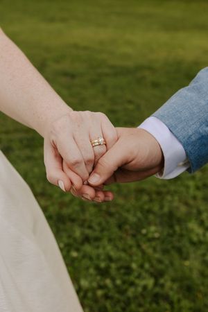 Close-up of bride and groom holding hands, bride wearing a gold wedding ring set on green grass background.