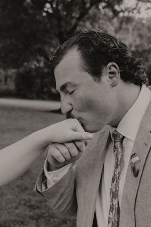 Groom in a light suit kisses bride's hand tenderly during outdoor wedding ceremony.