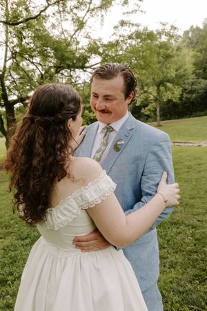 Smiling groom with mustache in blue suit embraces bride in white off-shoulder wedding dress outdoors.