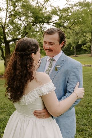 Groom in light blue suit laughs joyfully while embracing bride in white off-shoulder gown in a lush outdoor setting.