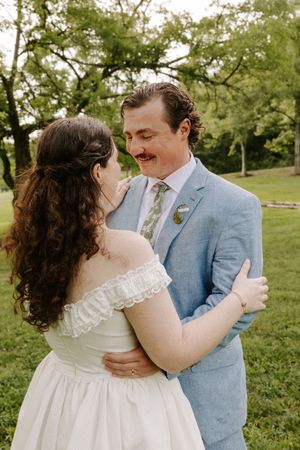 Bride and groom share a tender embrace outdoors, groom in light blue suit with floral tie smiling at his bride.