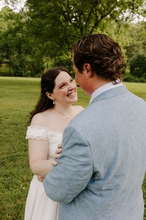 Bride laughing joyfully while holding grooms arm outdoors, wearing white off-shoulder gown and pearl earrings.