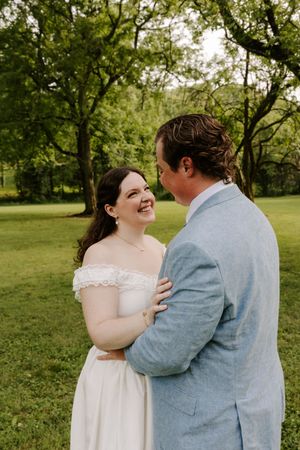 Bride laughs joyfully while embracing groom outdoors in green park setting during wedding portrait session.