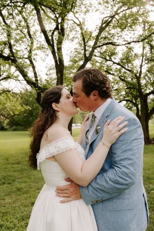 Bride and groom sharing a romantic kiss under large trees in a green garden on their wedding day.