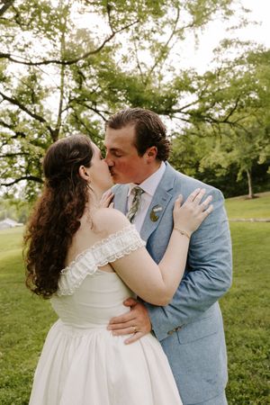 Bride and groom share a romantic kiss outdoors surrounded by lush green trees at their wedding.