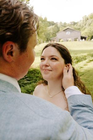 Groom in light blue suit tenderly brushes brides hair back as she smiles up at him near a barn venue.