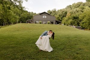 Groom dips bride for a romantic kiss on a green hillside with a dark barn wedding venue in the background.