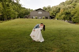 Groom dips bride for a kiss on a green hillside with a dark barn wedding venue visible in the background.