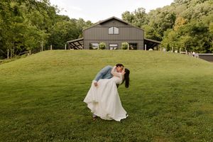 Groom dips bride in a romantic kiss in front of a modern gray barn wedding venue on a green hill.