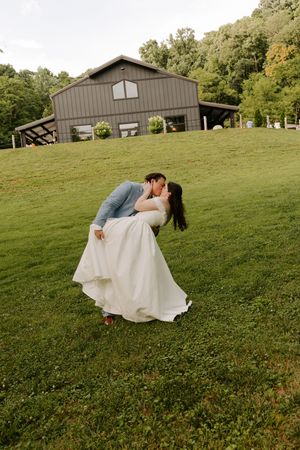 Groom dips bride for a romantic kiss on a green hillside with a charming dark barn wedding venue in the background.