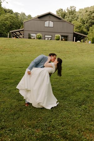 Groom dips bride for a romantic kiss on green lawn with a gray barn wedding venue in background.