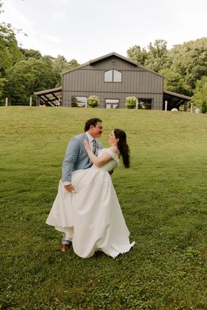 A groom dips his bride for a kiss on a green hillside with a rustic barn venue in the background.