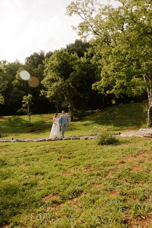 Bride and groom walk hand-in-hand through a scenic outdoor wedding venue surrounded by lush green trees and landscape.