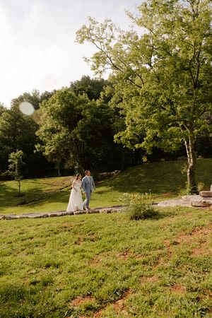 A bride and groom walk hand in hand through a lush green outdoor venue on their wedding day.