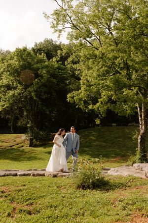 Bride and groom hold hands while walking on stone steps surrounded by lush green trees outdoors.
