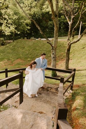 Groom helps bride carry her white gown up stone steps on a wooden bridge during wedding portraits.
