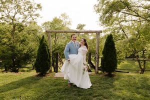 Smiling bride and groom walk together beneath a rustic wooden arbor flanked by evergreen trees at outdoor wedding venue.