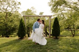 Newlyweds share a kiss under a rustic wooden arch surrounded by evergreen trees at outdoor wedding.