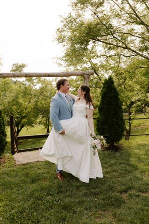 Bride and groom share a joyful moment on garden lawn with rustic wooden fence and tall trees behind them.