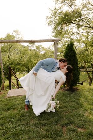 Groom dramatically dips bride for a kiss near a rustic wooden arbor with evergreen trees at an outdoor wedding venue.