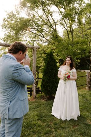 Groom in blue suit photographs bride in white off-shoulder gown holding bouquet outdoors at wedding.